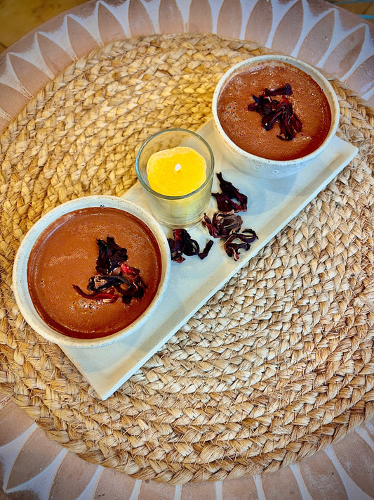 Momento íntimo con dos tazas de cacao infusionado y una vela encendida sobre mesa rústica. Aroma a hibisco y madera, inspiración y calor interior.