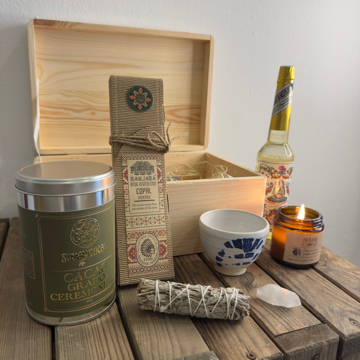 Caja de madera con los elementos del kit regalo King, lata cacao ceremonial, copal, agua florida, vela, taza, cuarzo, salvia, sobre mesa de madera.