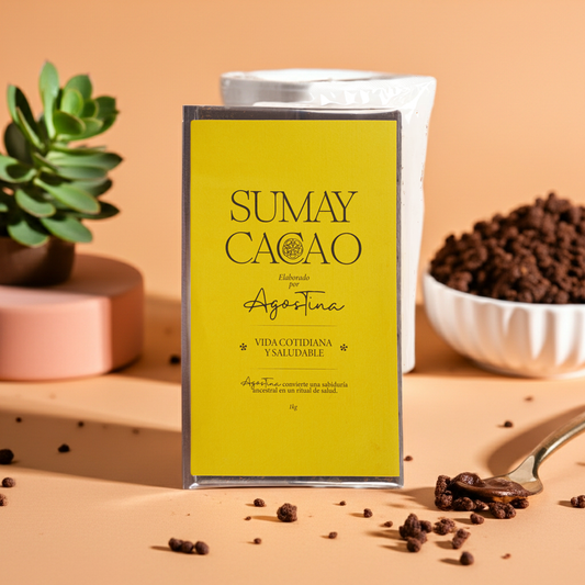 Pasta de cacao Vida Cotidiana de Sumay Cacao, bloque de 1 kg con etiqueta amarilla sobre fondo melocotón, acompañado de cacao triturado y una planta suculenta