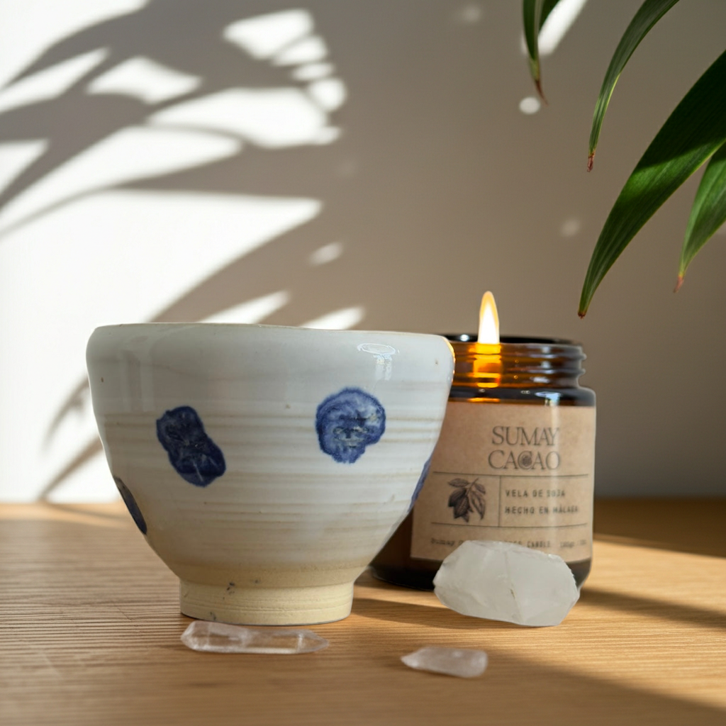 Tacita artesanal de cerámica blanca con pintitas azules sobre mesa de madera clara, junto a una vela encendida y cristales de cuarzo, con sombras de hojas al fondo.