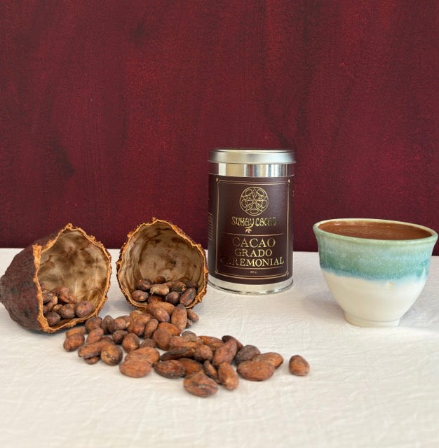 Lata de cacao ceremonial Sumay con taza de cacao sobre mesa blanca con granos de cacao saliendo de una cáscara seca