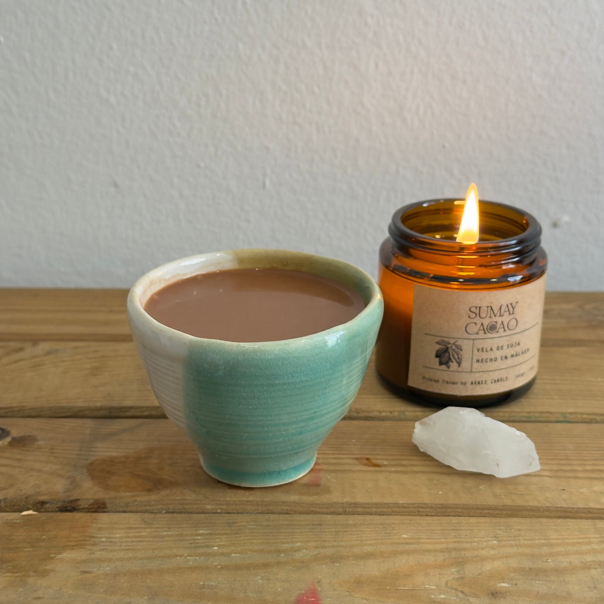 Taza artesanal con cacao ceremonial, cuarzo y vela con pared blanca de fondo.