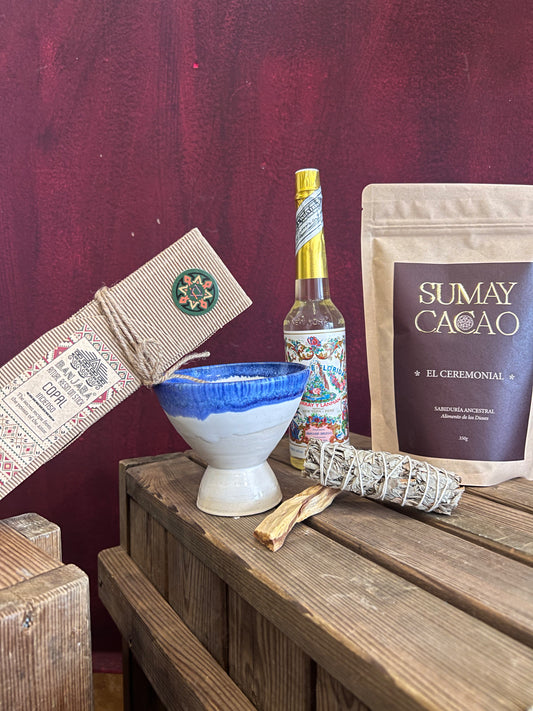 Paquete de cacao ceremonial y accesorios rituales (agua florida, salvia, copal, palo santo y copalera) sobre cajón de madera con fondo rojo oscuro.