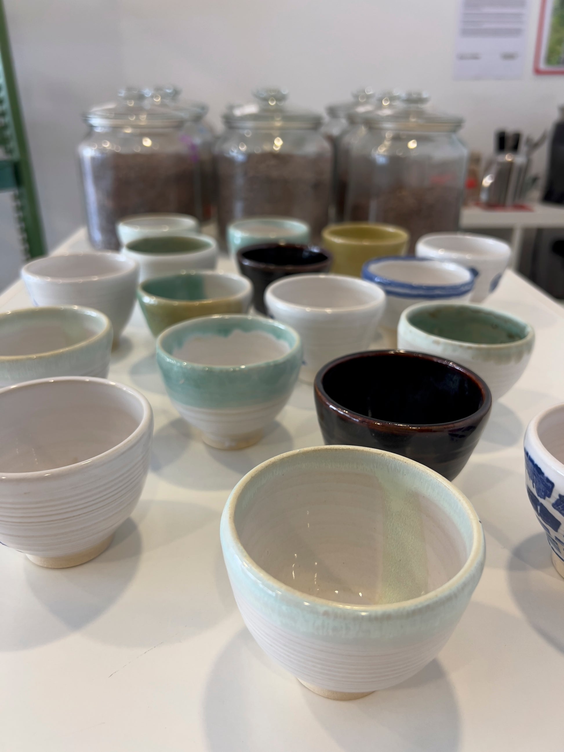 Diferentes diseños de tazas de cerámica artesanal sobre mesa blanca y fondo con frascos de cacao.