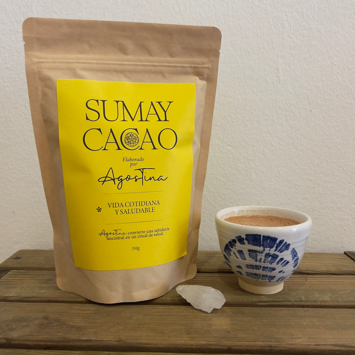 Paquete de 350g Sumay Cacao con cuarzo y taza de cerámica sobre mesa madera y fondo blanco.